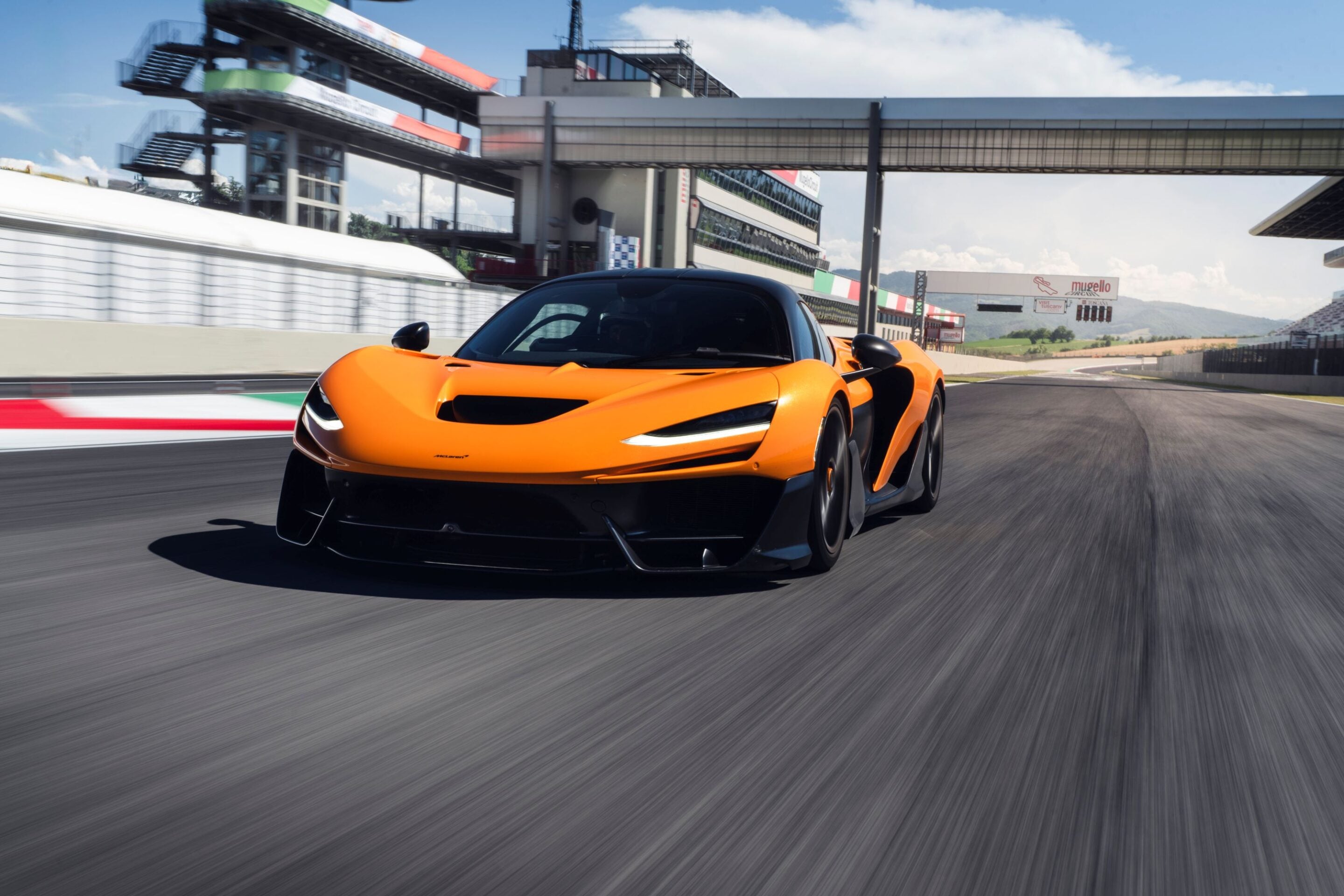 Orange McLaren sports car speeding on a race track with pit buildings and grandstands in the background under a blue sky.