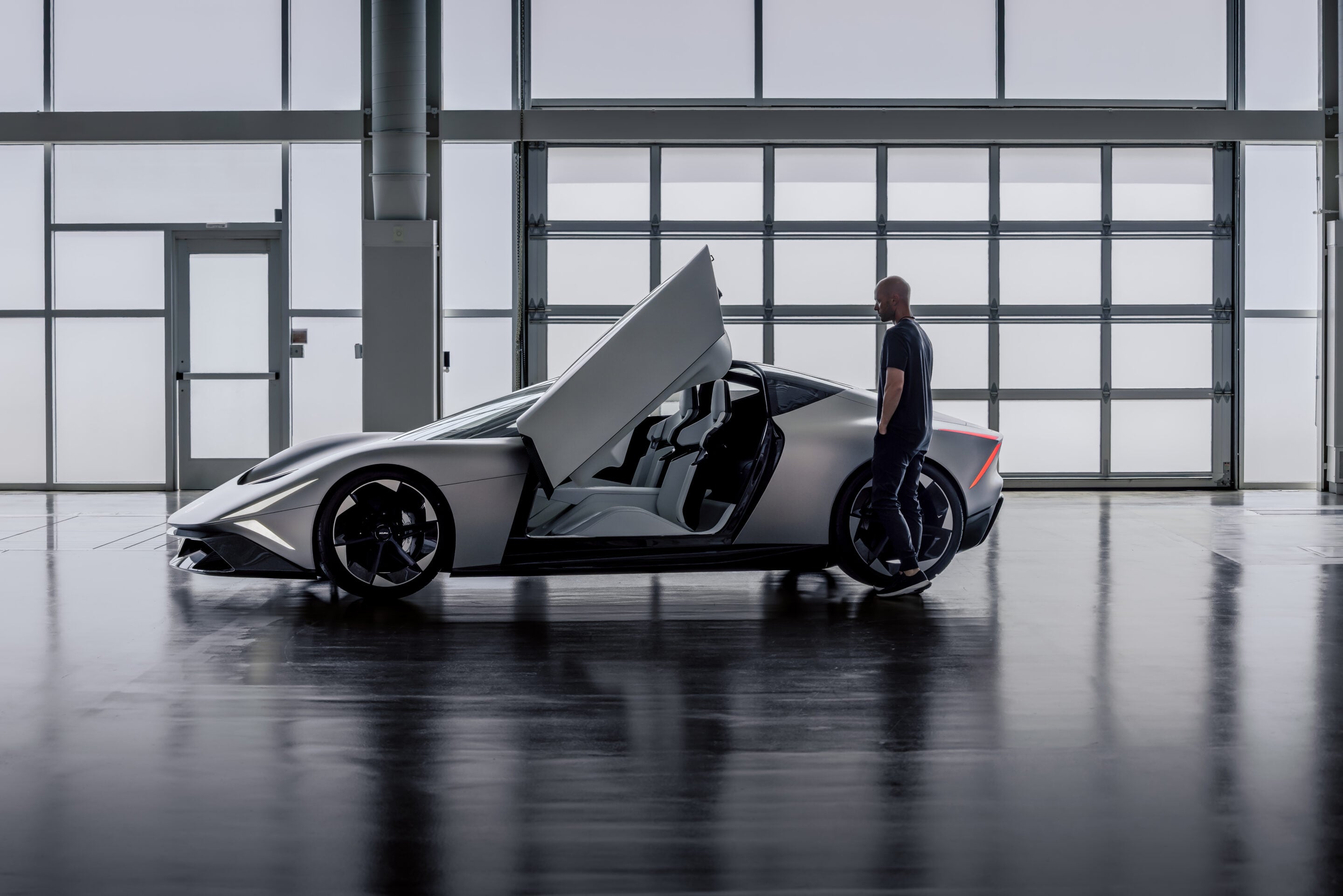 Man in a dark shirt stands beside a white futuristic sports car with upward-opening doors inside a bright showroom or hangar.