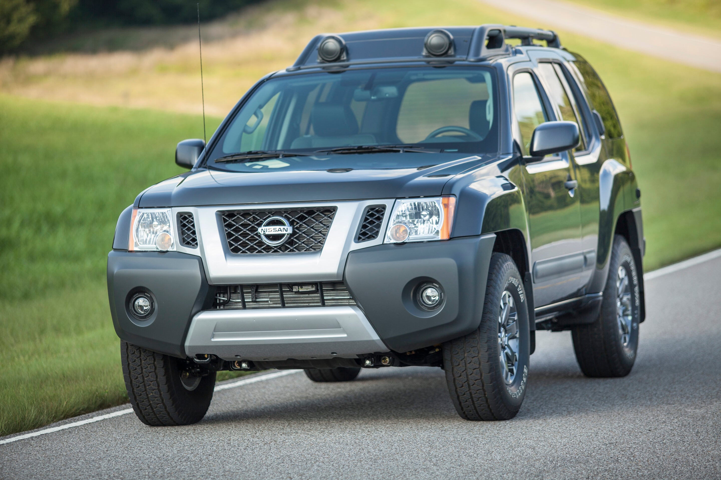 Front-left view of a gray Nissan SUV driving on a rural road with green fields beside it.