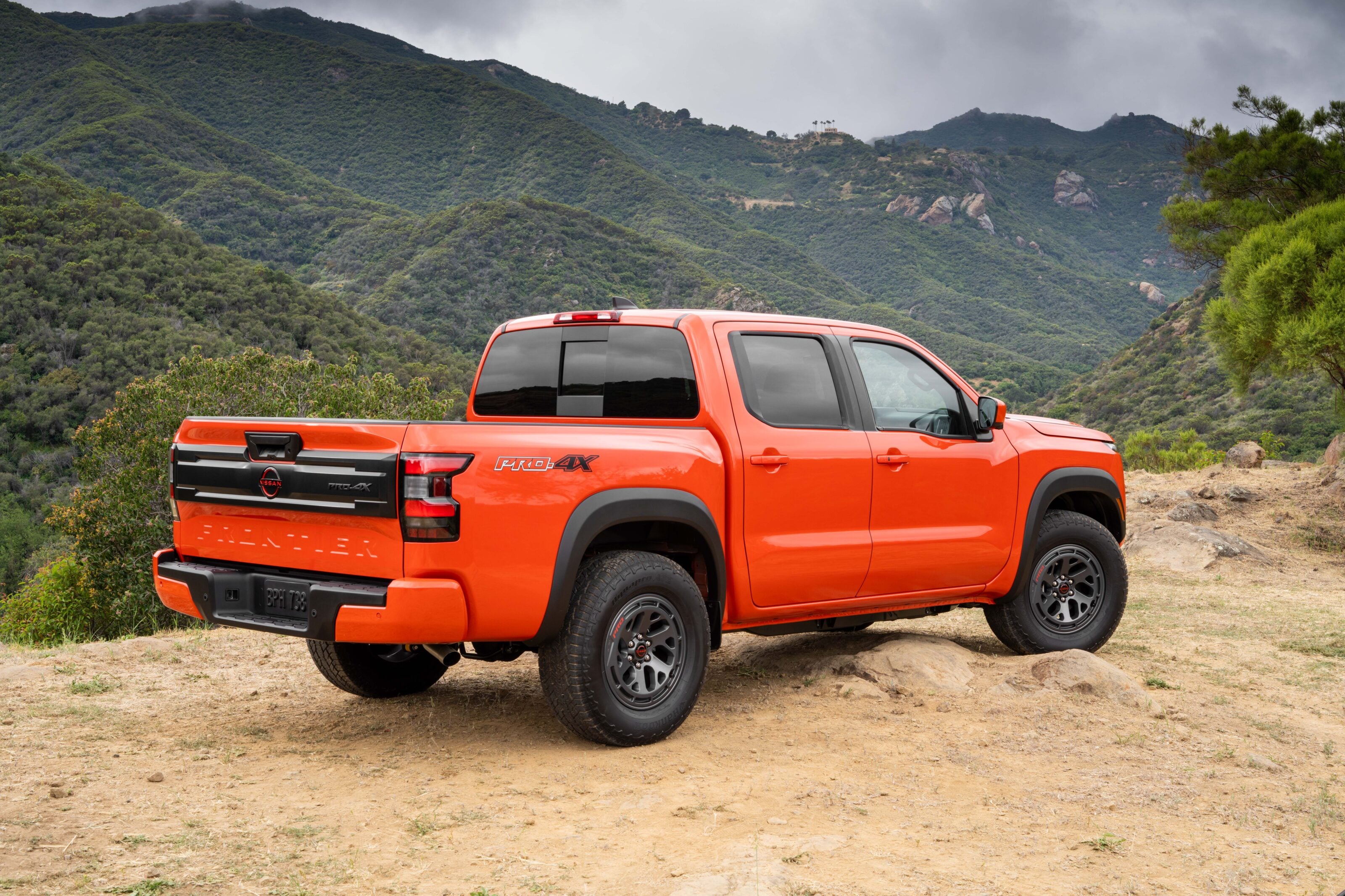 Orange Nissan Pro-4X pickup parked on a dirt trail with green mountains and a cloudy sky in the background.