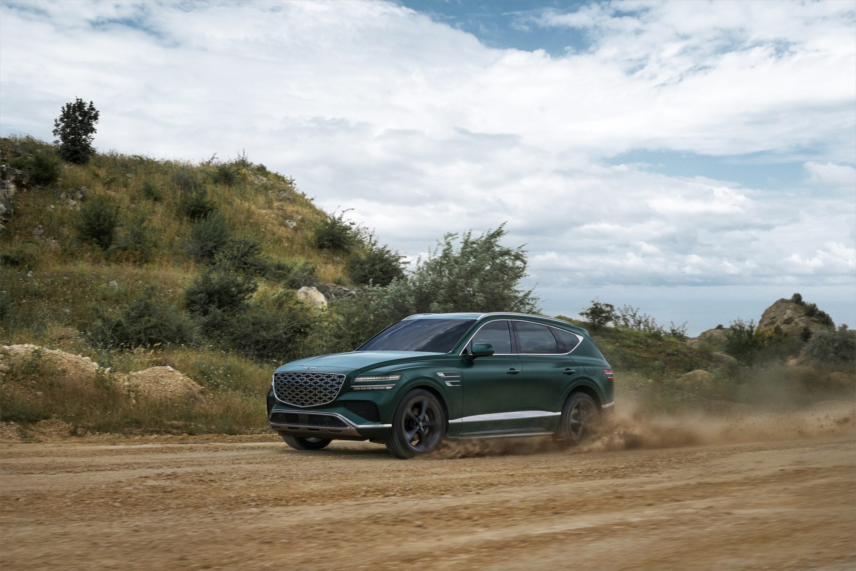 Green SUV driving on a dirt road with grassy hills and a cloudy sky in the background.