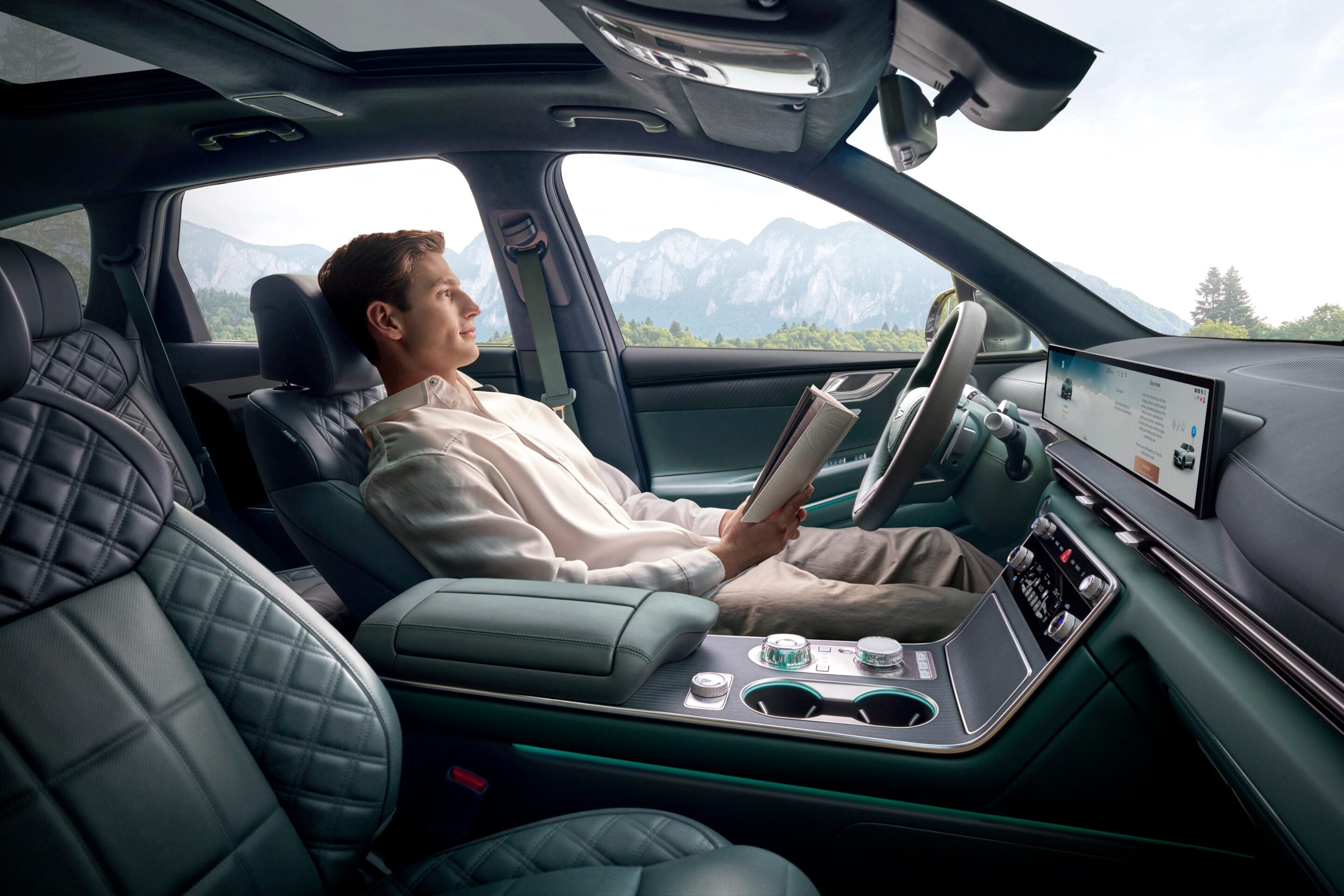 Man seated in the left front passenger seat of a luxury car, reading a magazine as a mountain landscape passes by the window.