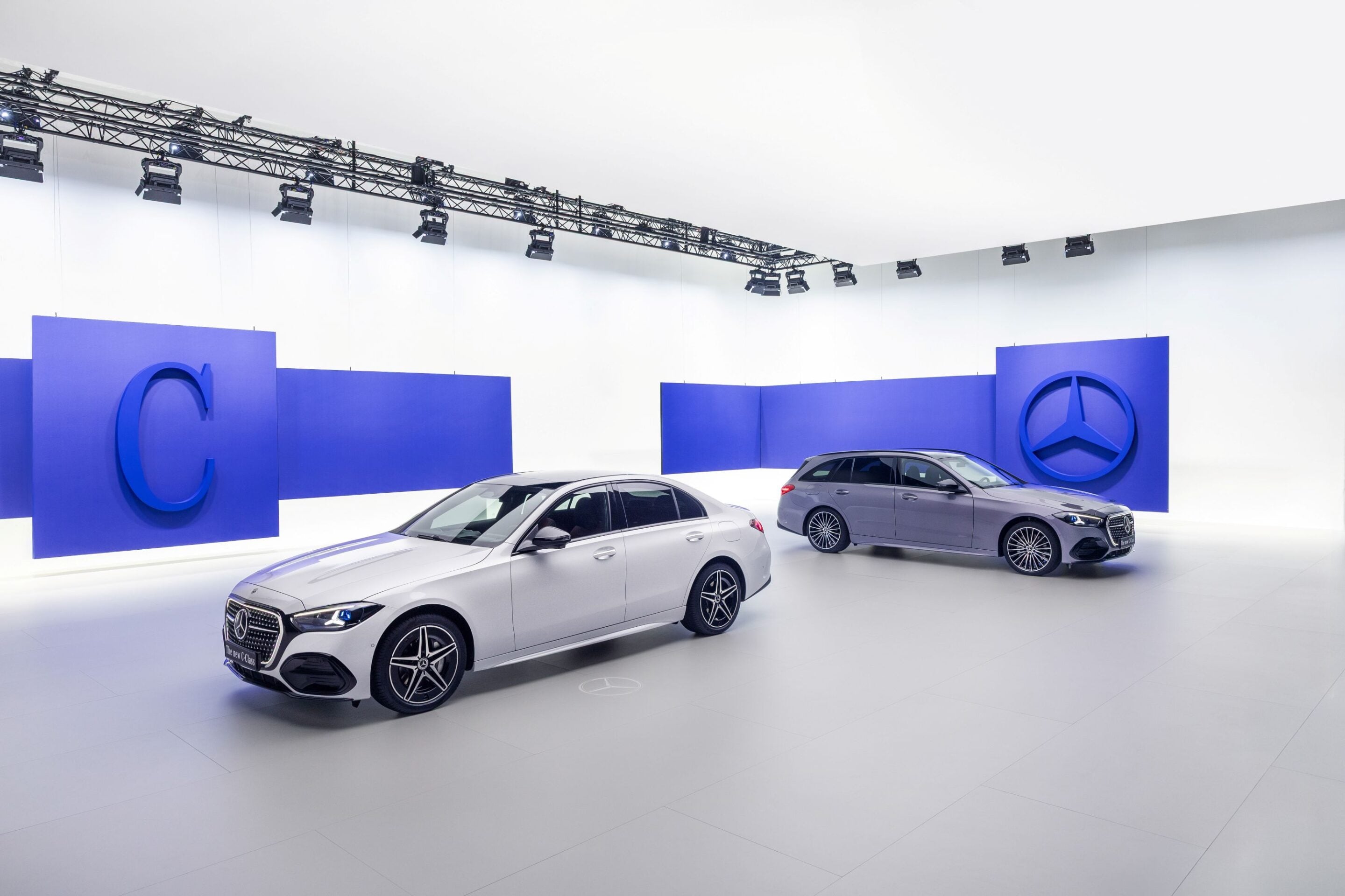 Mercedes-Benz cars on display in a bright showroom with blue wall panels and a large Mercedes logo backdrop.