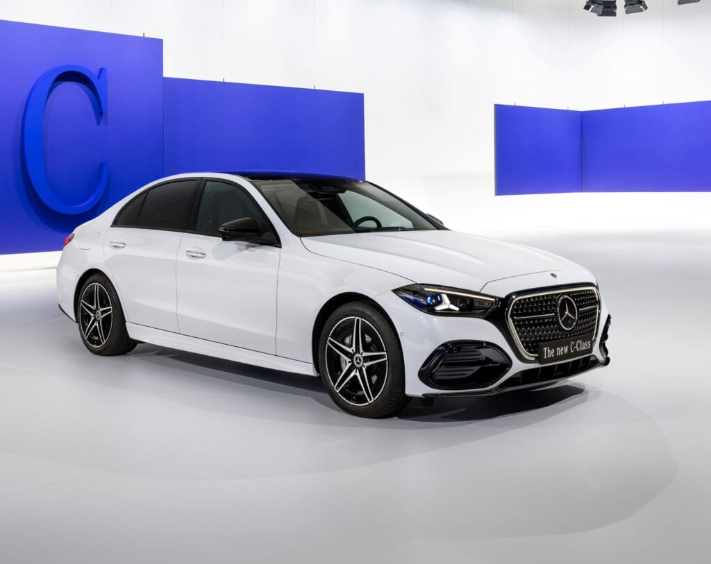 White Mercedes-Benz C-Class sedan on a glossy showroom floor, blue wall panels in the background, plate reads 'The new C-Class'