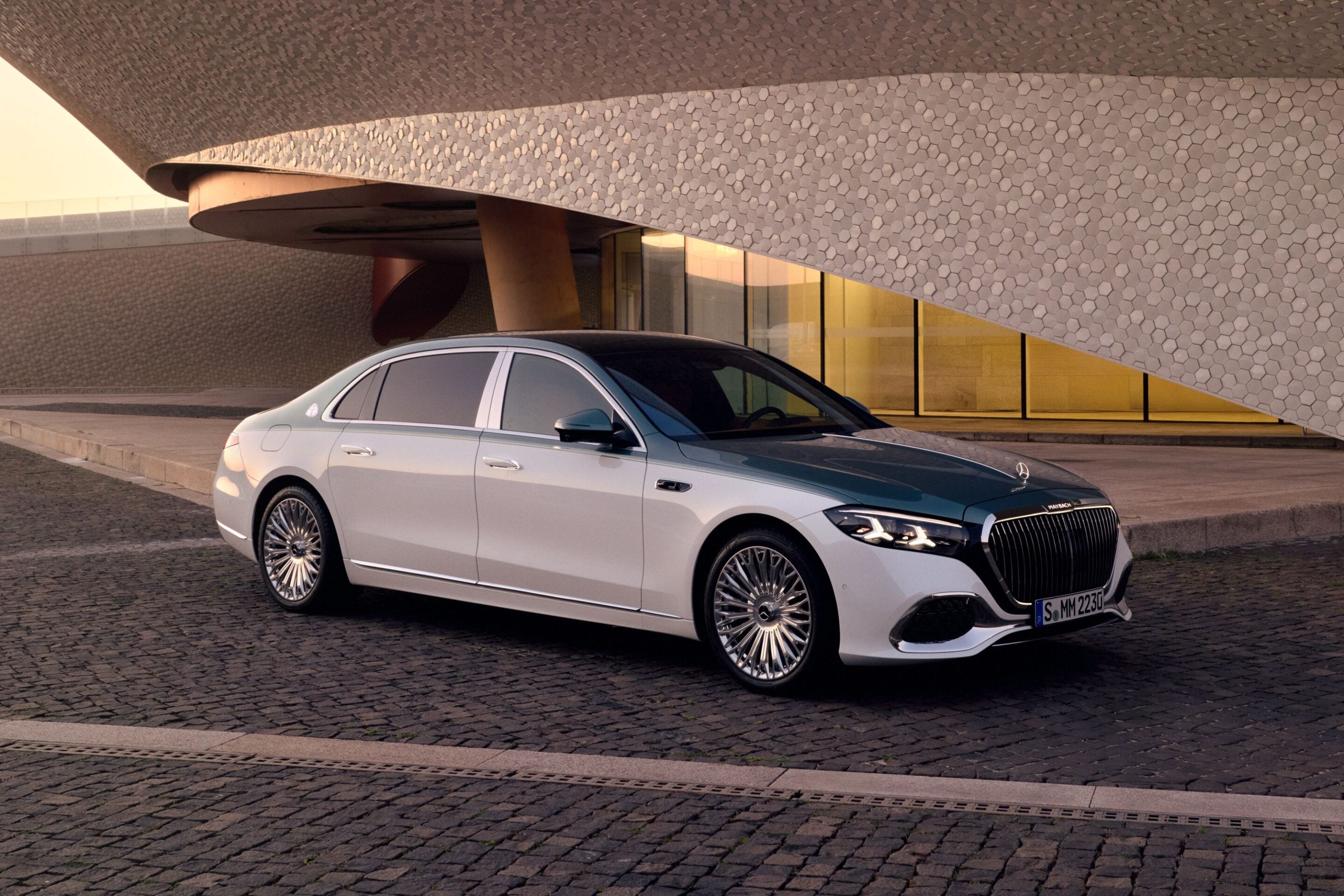 Silver luxury Mercedes-Maybach sedan parked on a cobblestone street in front of a modern honeycomb-patterned building.
