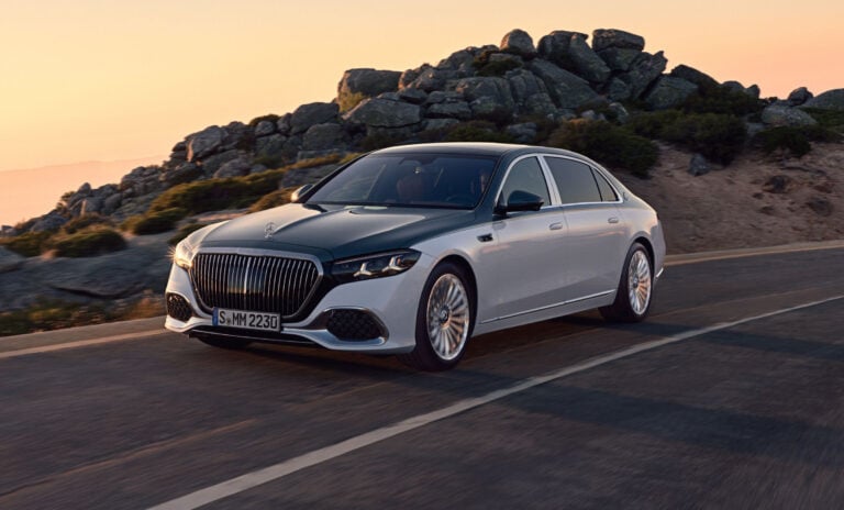 Front three-quarter view of a silver Mercedes sedan driving on a winding coastal road at sunset with rocky hills in the background.