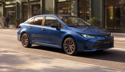 Blue Toyota Corolla Hybrid sedan on a city street, front three-quarter view at curb level edge of a sidewalk.