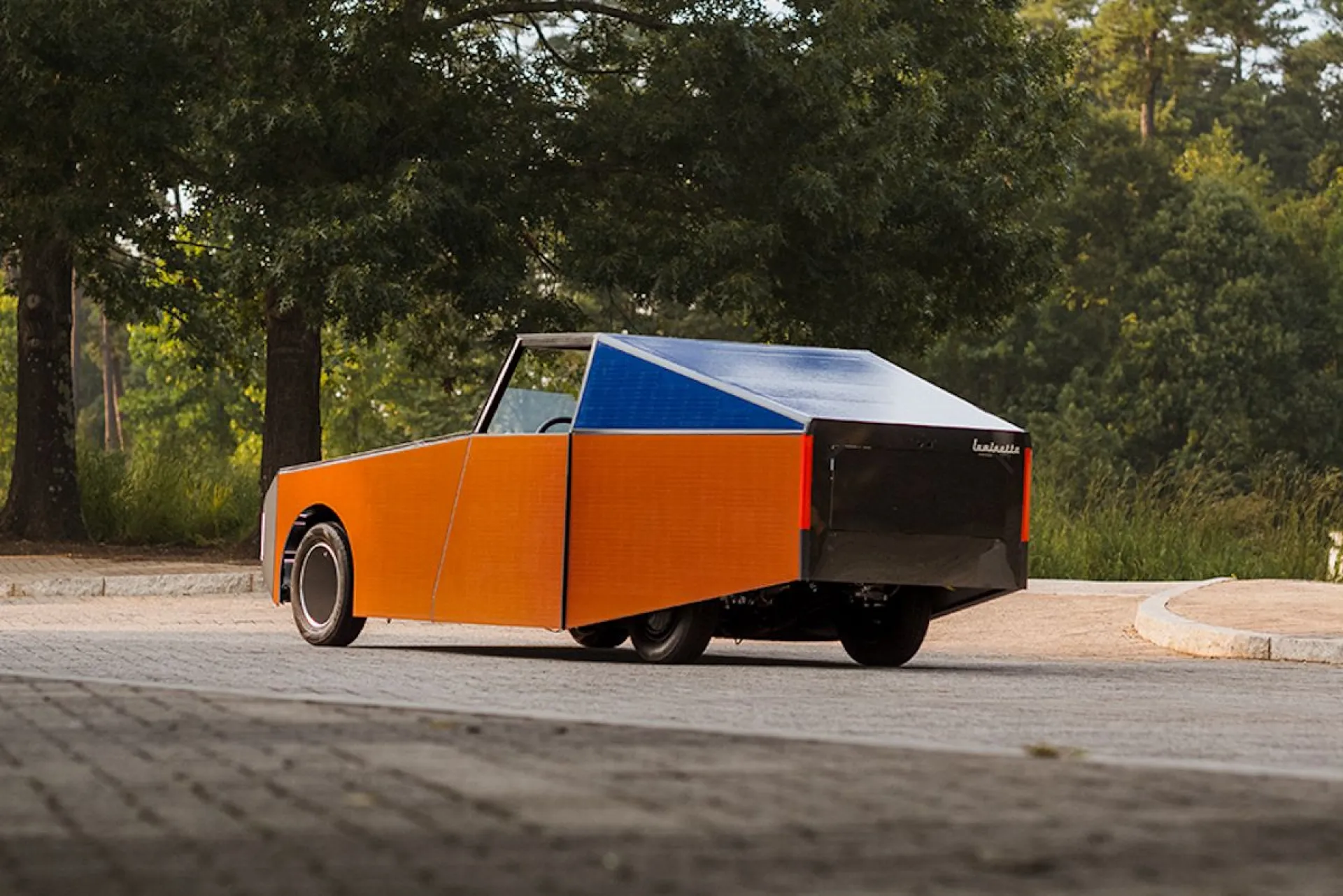Orange, boxy concept car with a blue roof parked on a paved street near trees in a park-like setting.