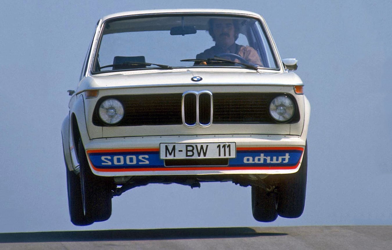 Front view of a white vintage BMW jumping off a ramp; license plate M-BW 111; driver visible in the cockpit against a blue sky.