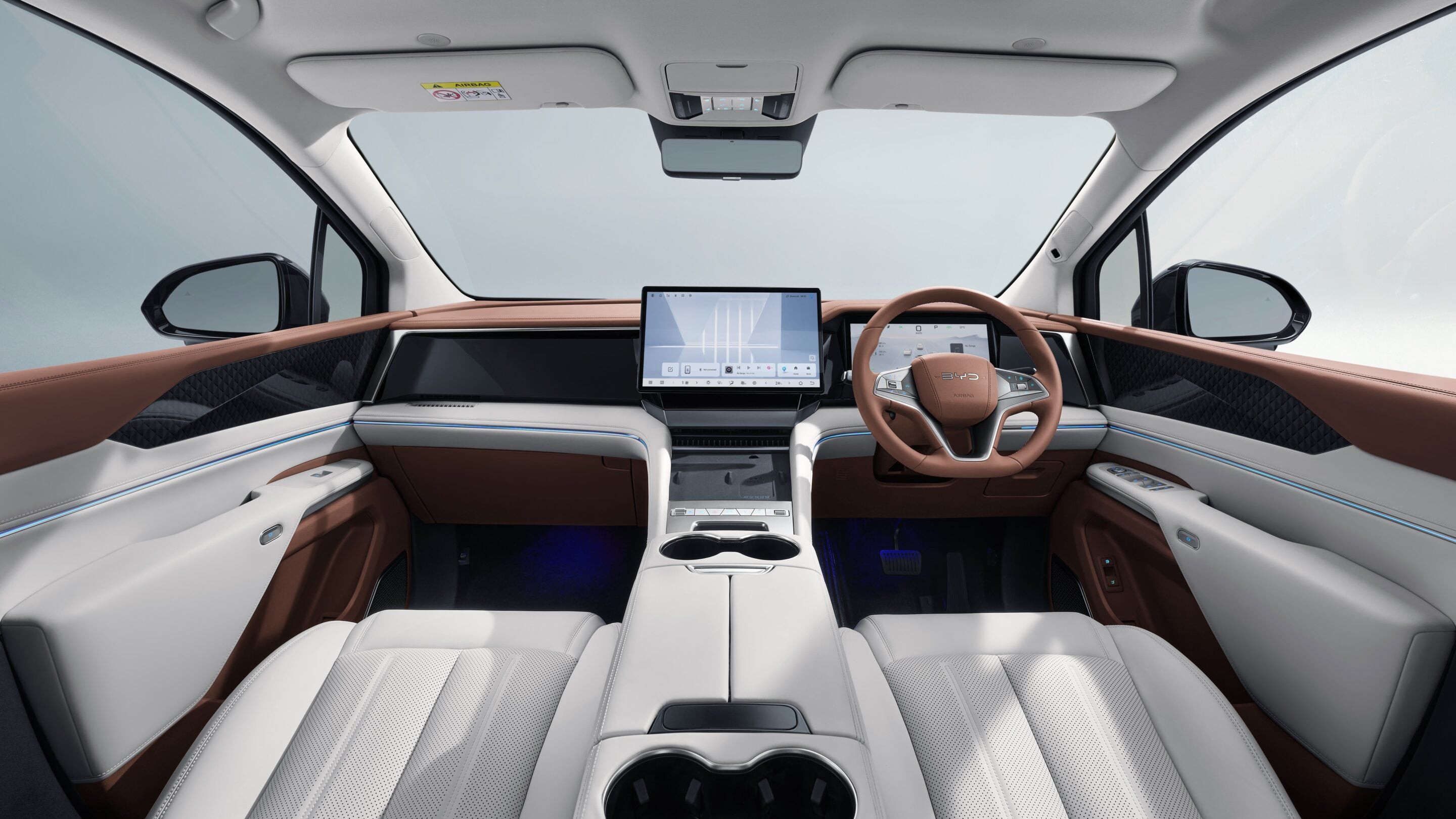 Luxurious car interior from rear passenger perspective with tan leather seats and a wide digital dashboard including a large center touchscreen and steering wheel.