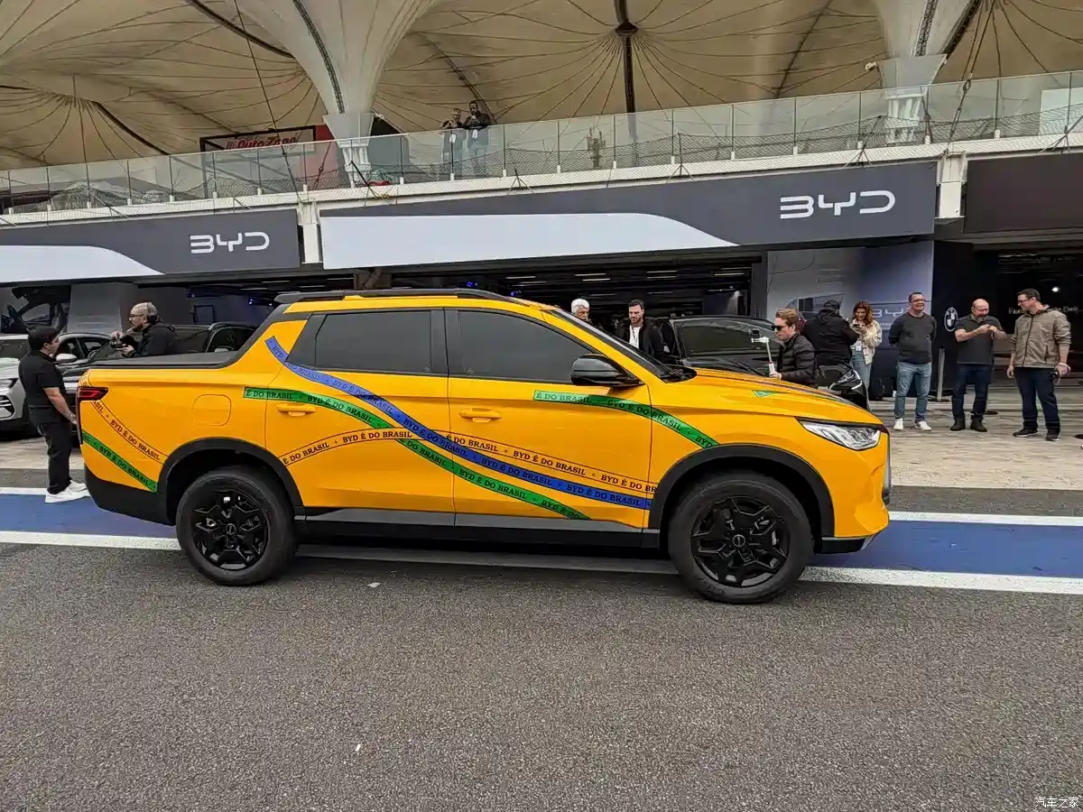 Orange BYD SUV/pickup with green and blue tape reading 'BYD é do Brasil' diagonally across the body, shown at a car event with bystanders in the background.
