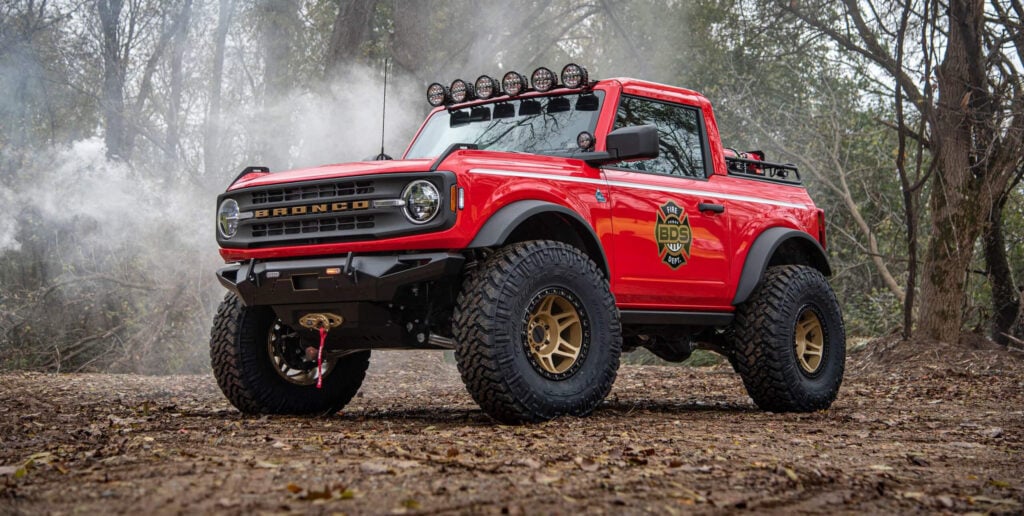 Red Ford Bronco off-road truck with large tires and roof lights in a smoky forest backdrop, showing a Fire Dept badge on the door.