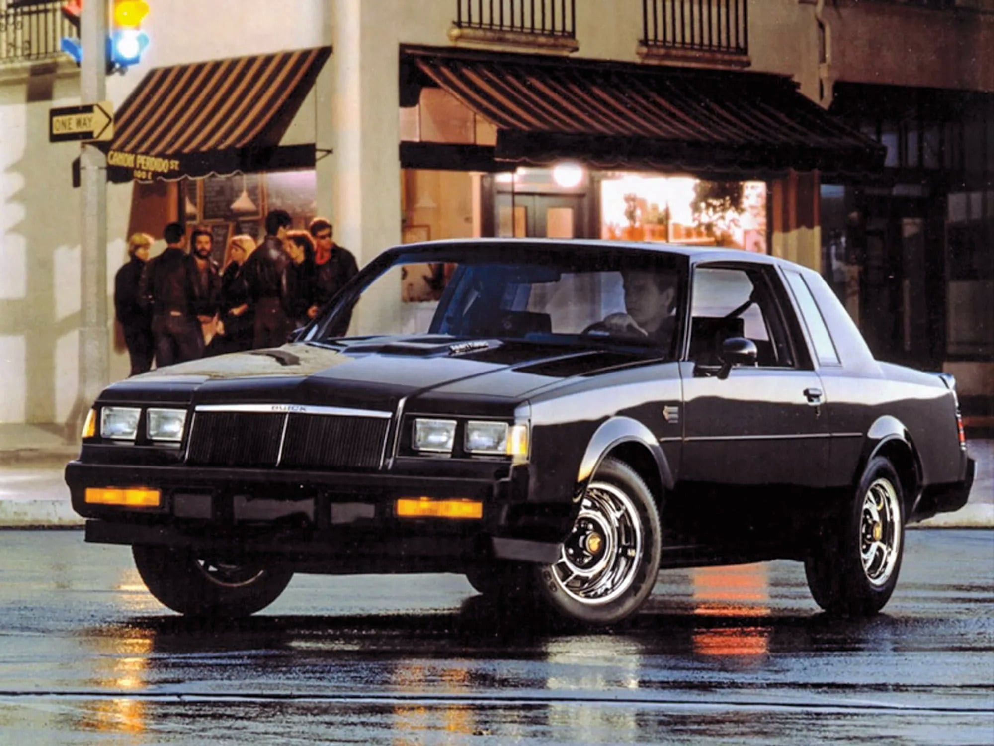 Front three-quarter view of a black SUV parked on a rain-wet street at night, with storefronts and pedestrians in the background.
