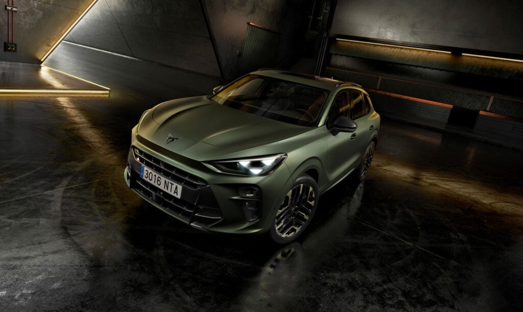 Front three-quarter view of a matte olive-green SUV with bold grille and alloy wheels in a dim, industrial garage lit by warm strip lights along the walls.
