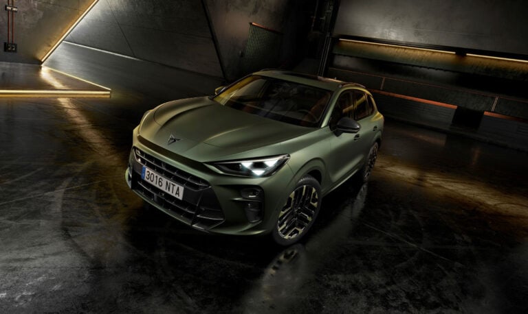 Front three-quarter view of a matte olive-green SUV with bold grille and alloy wheels in a dim, industrial garage lit by warm strip lights along the walls.