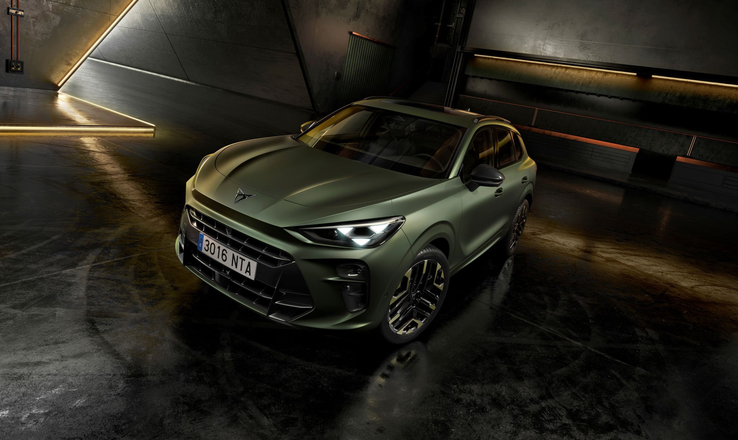 Front three-quarter view of a matte olive-green SUV with bold grille and alloy wheels in a dim, industrial garage lit by warm strip lights along the walls.