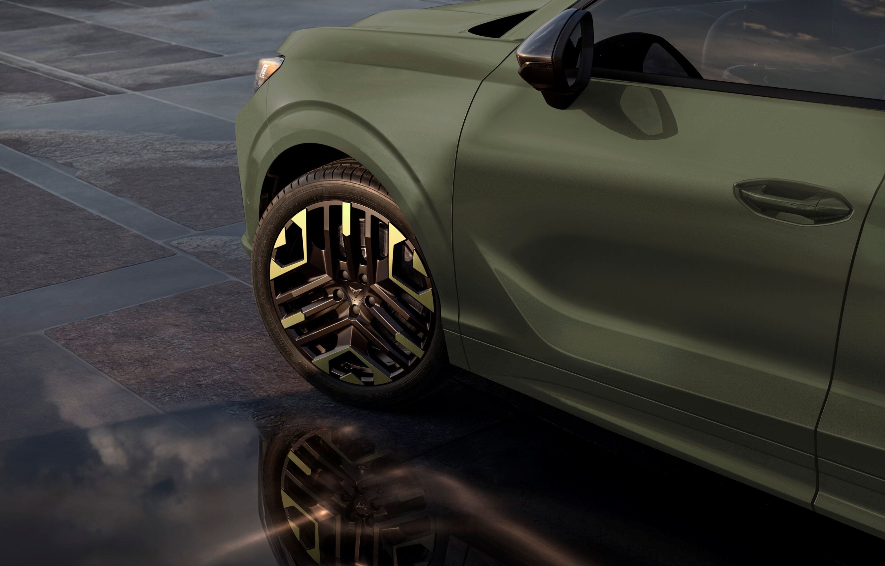 Close-up of a green SUV's front left wheel, fender, and door on a paved surface with a reflective body panel in view of the side mirror.