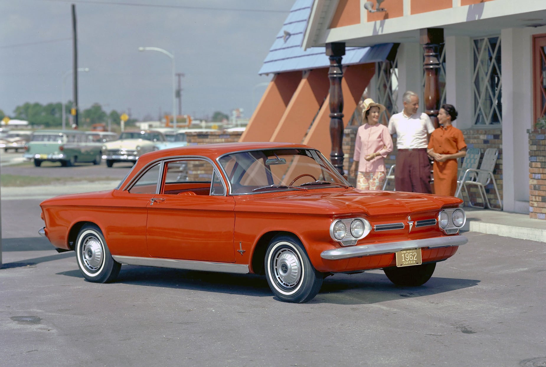 Red vintage car parked in front of a mid-century motel while three adults chat near the entrance.
