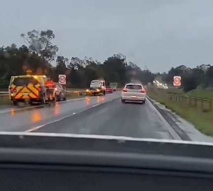 View from a car on a wet highway at dusk with several cars ahead stopped in traffic; a yellow service/emergency vehicle is on the shoulder with flashing lights.