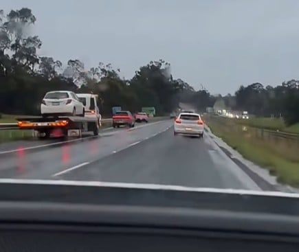 Wet highway with several cars; a tow truck on the left carrying a white SUV on a flatbed trailer.