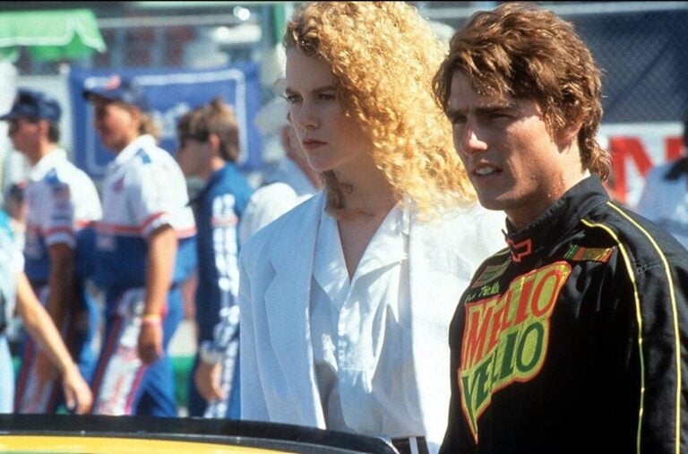 Two race crew members stand in a busy pit area, looking serious and focused on something off-camera.