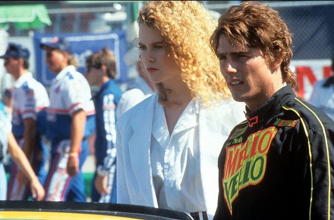 Two race crew members stand in a busy pit area, looking serious and focused on something off-camera.