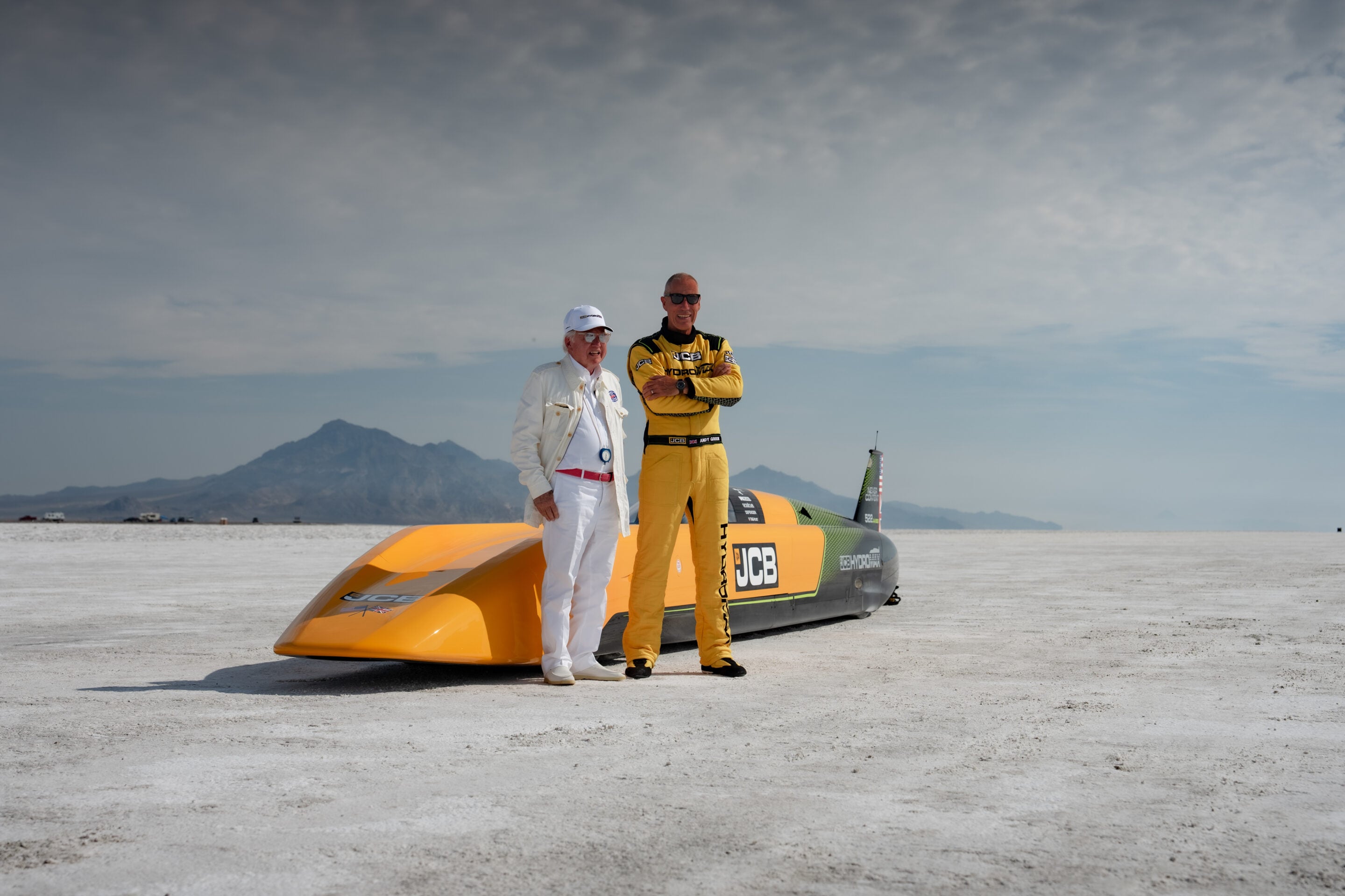 Two men stand beside an orange speedboat on a salt flat with distant mountains under a clear sky.