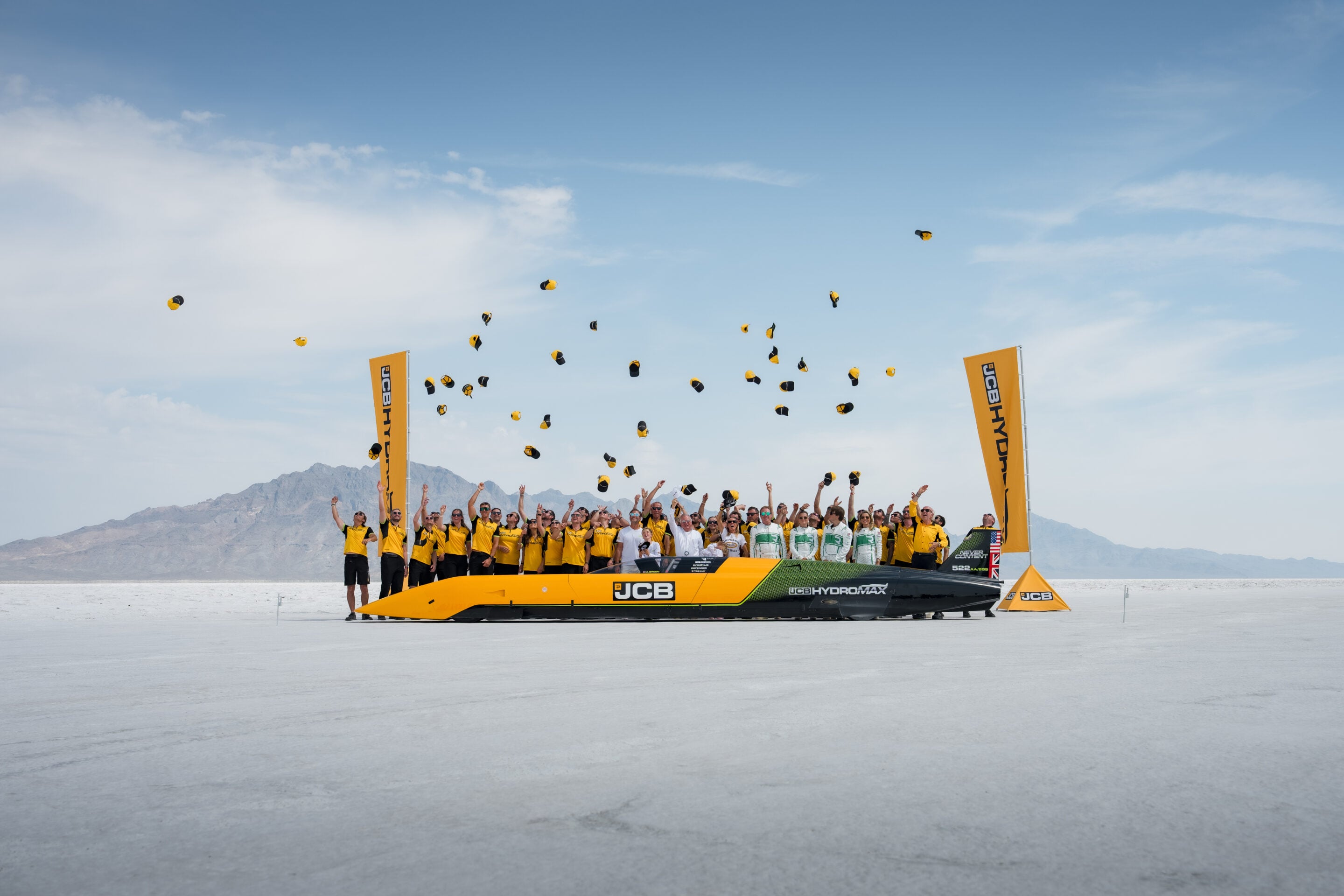 Crowd of people in yellow shirts celebrating around a bright yellow JCB HydroMAX speedboat on a white salt flat, tossing yellow hats into the air under a blue sky.