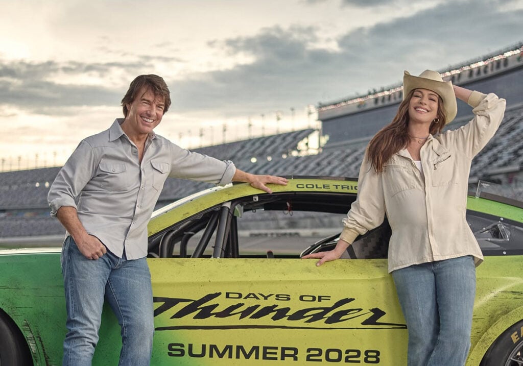 Smiling man and woman in casual clothing pose with a lime-green race car at a racetrack, the car showing 'Days of Thunder' and 'Summer 2028' text on its side.