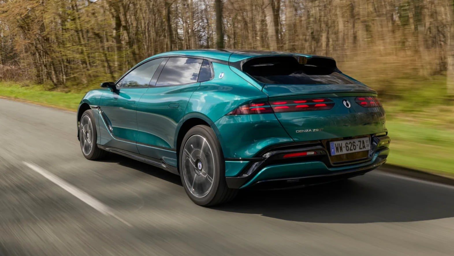 Teal BMW Denza SUV driving away on a countryside road, rear three-quarter view with motion blur behind it.