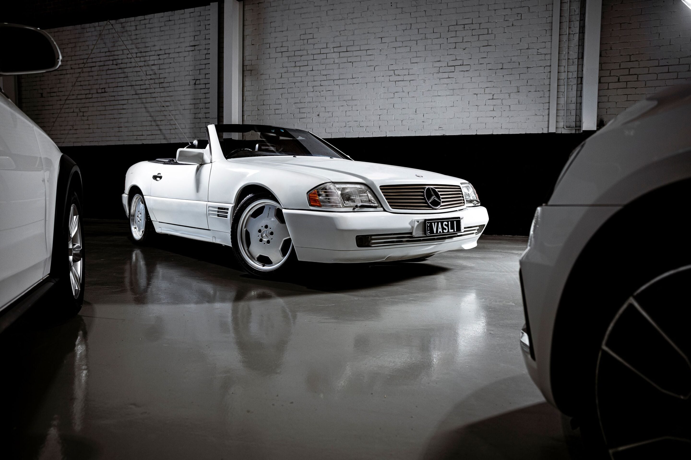 White Mercedes-Benz convertible parked in a dim indoor garage with a brick wall behind it and other cars nearby, viewed from a low angle to the front-left side.] ,