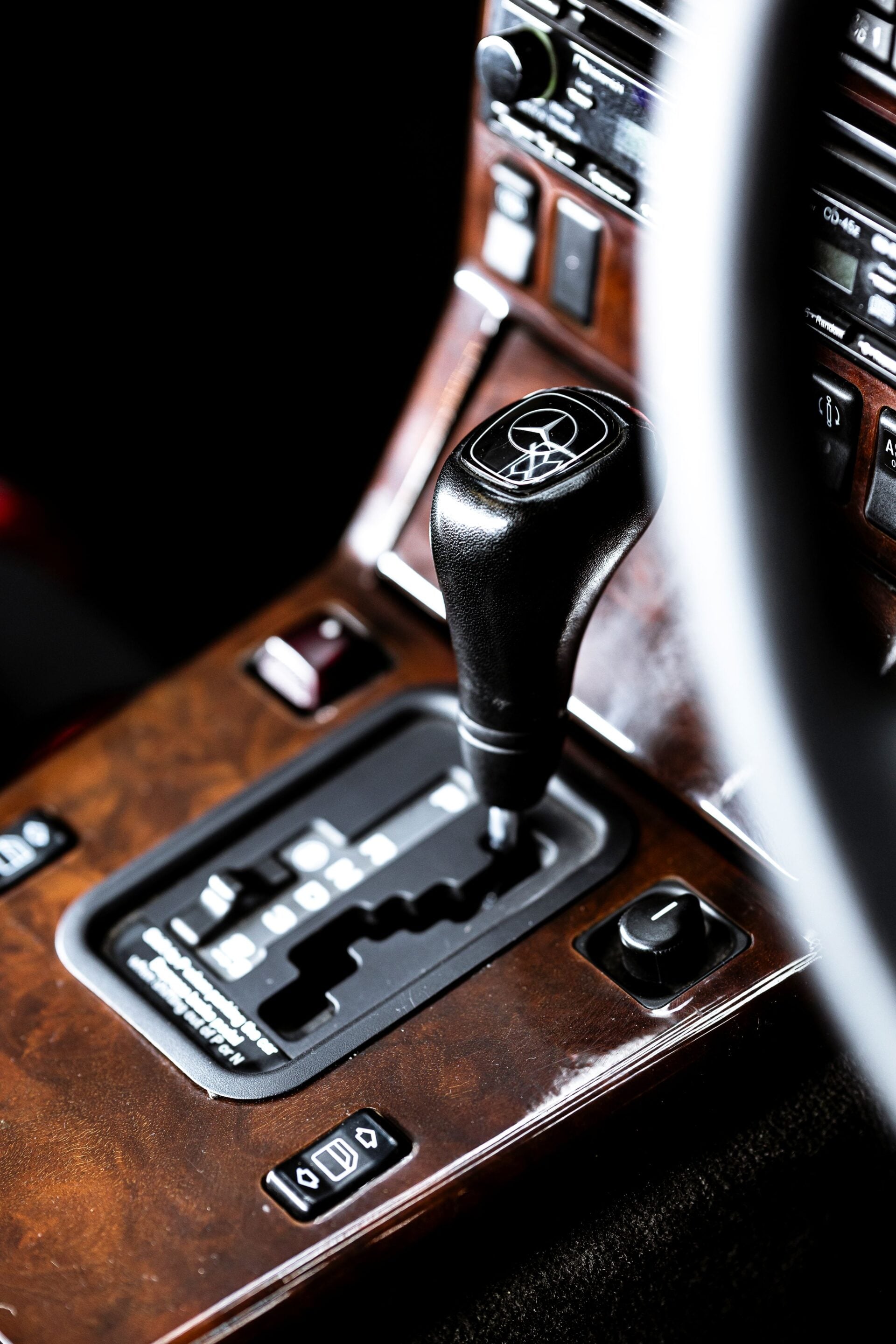 Close-up of a black gear shift knob with a Mercedes emblem, set on a brown wood-trimmed console inside a car.