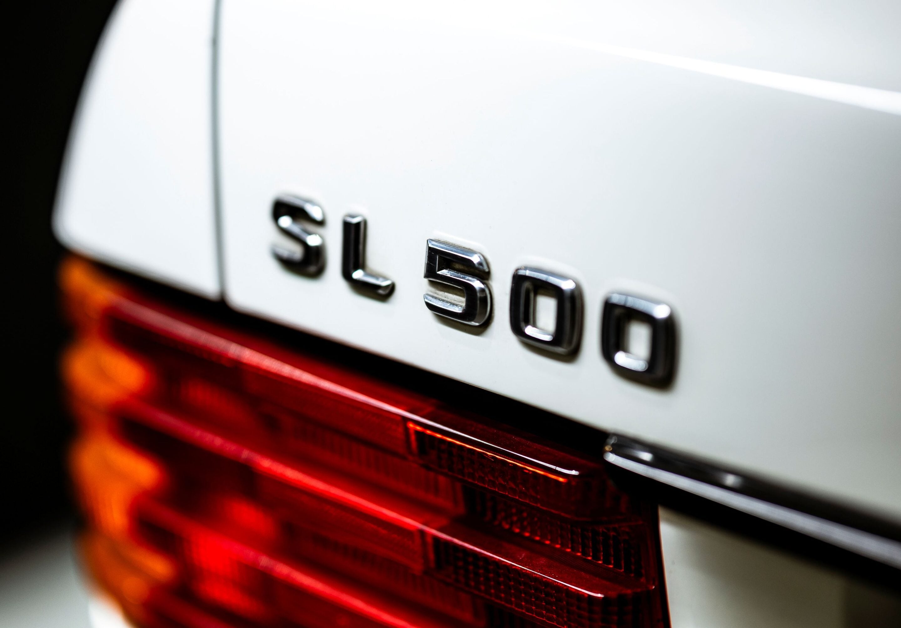 Close-up of a white car rear showing a chrome 'SL500' badge and red taillight, with shallow depth of field.