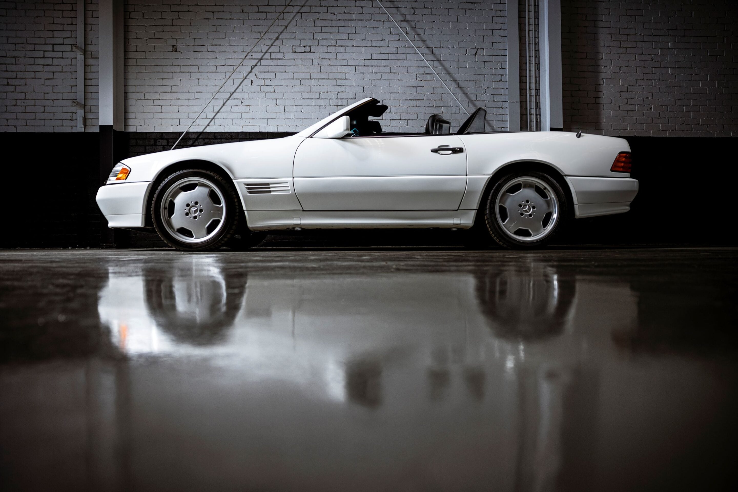 White convertible car parked indoors beside a brick wall, reflection visible on the glossy floor.