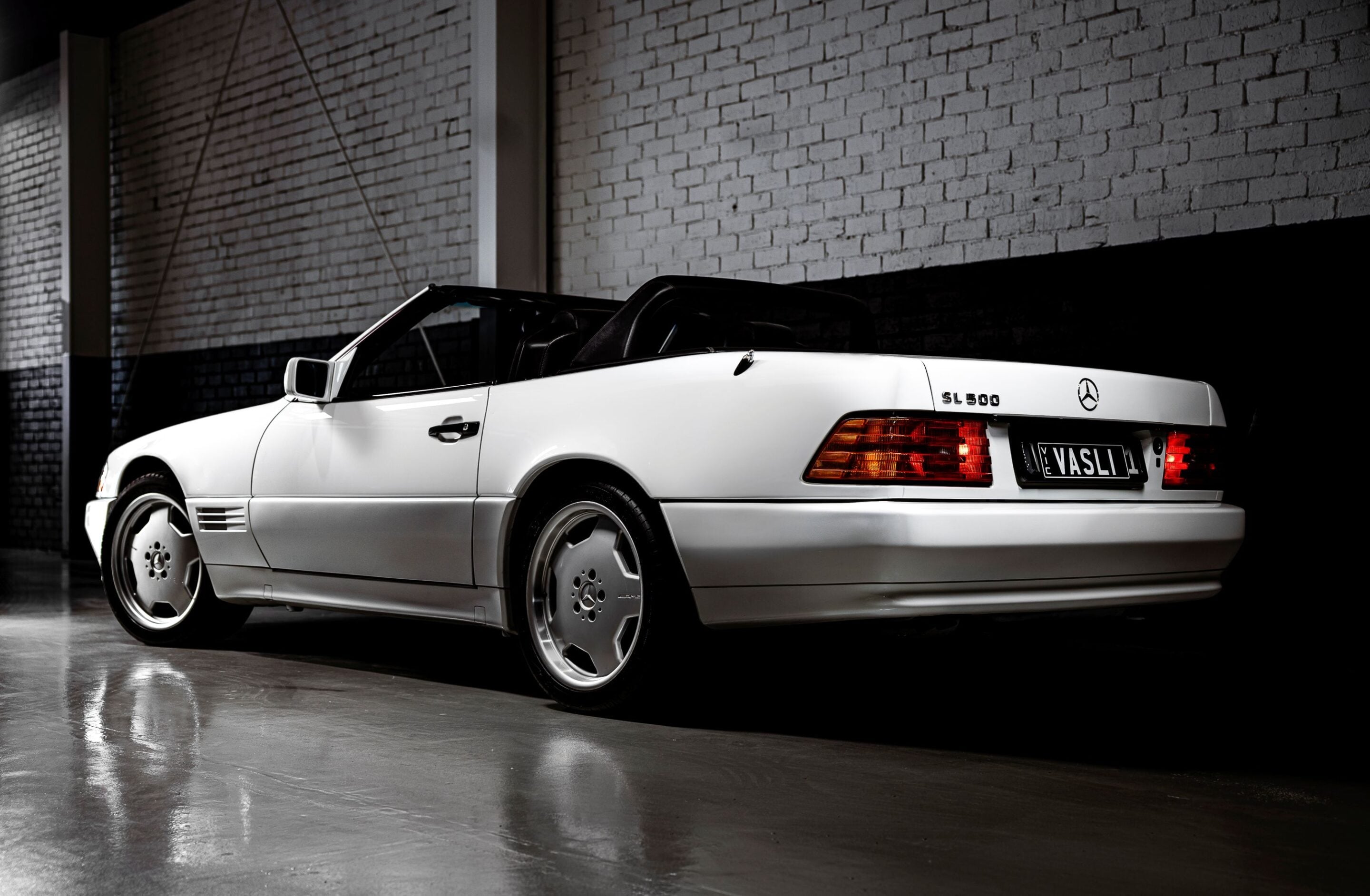 White Mercedes-Benz SL500 convertible parked in a dim garage with brick walls in the background, rear quarter view.