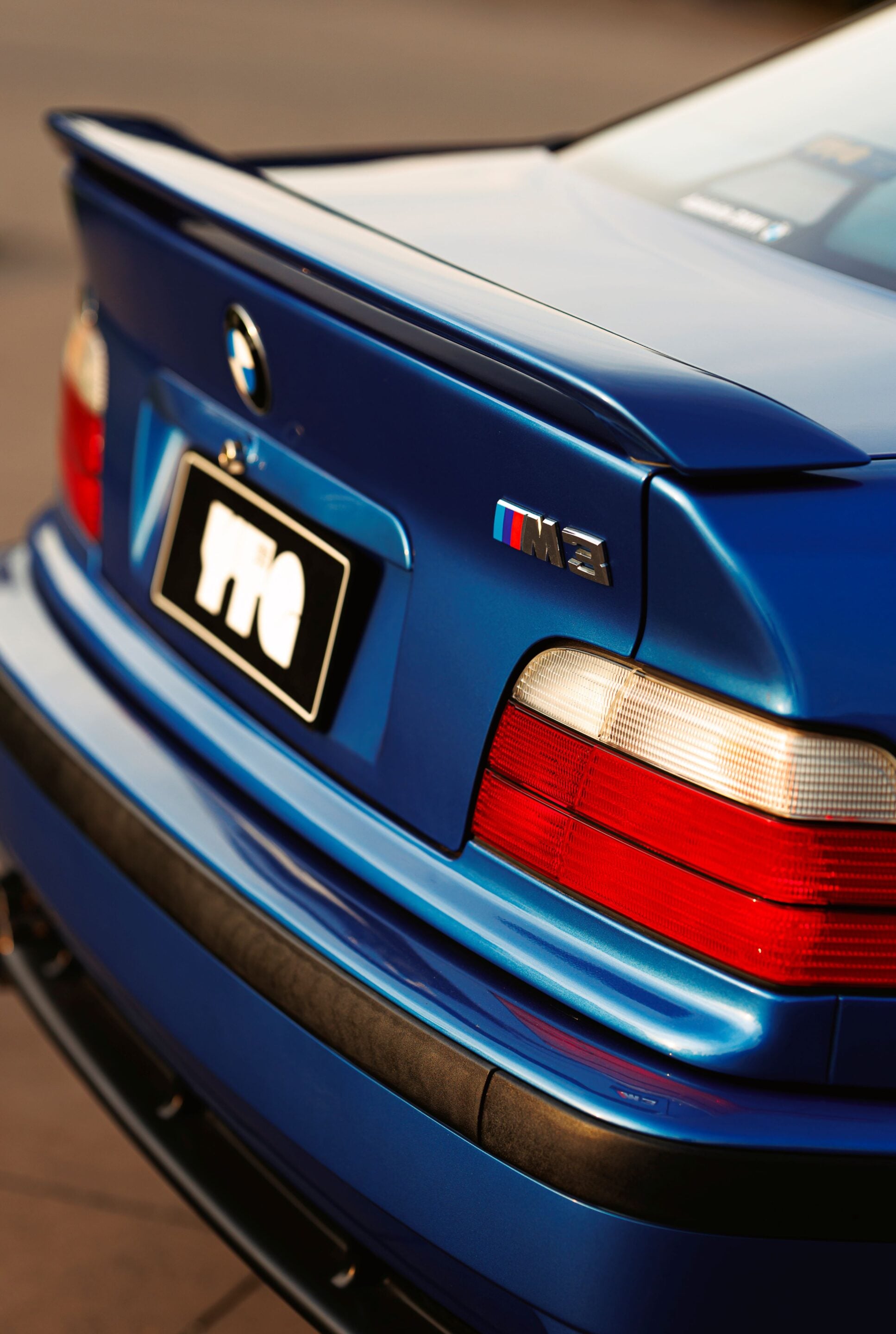Blue BMW M3 rear with trunk spoiler and prominent taillight cluster, close-up detail of the badge and bumper edge.