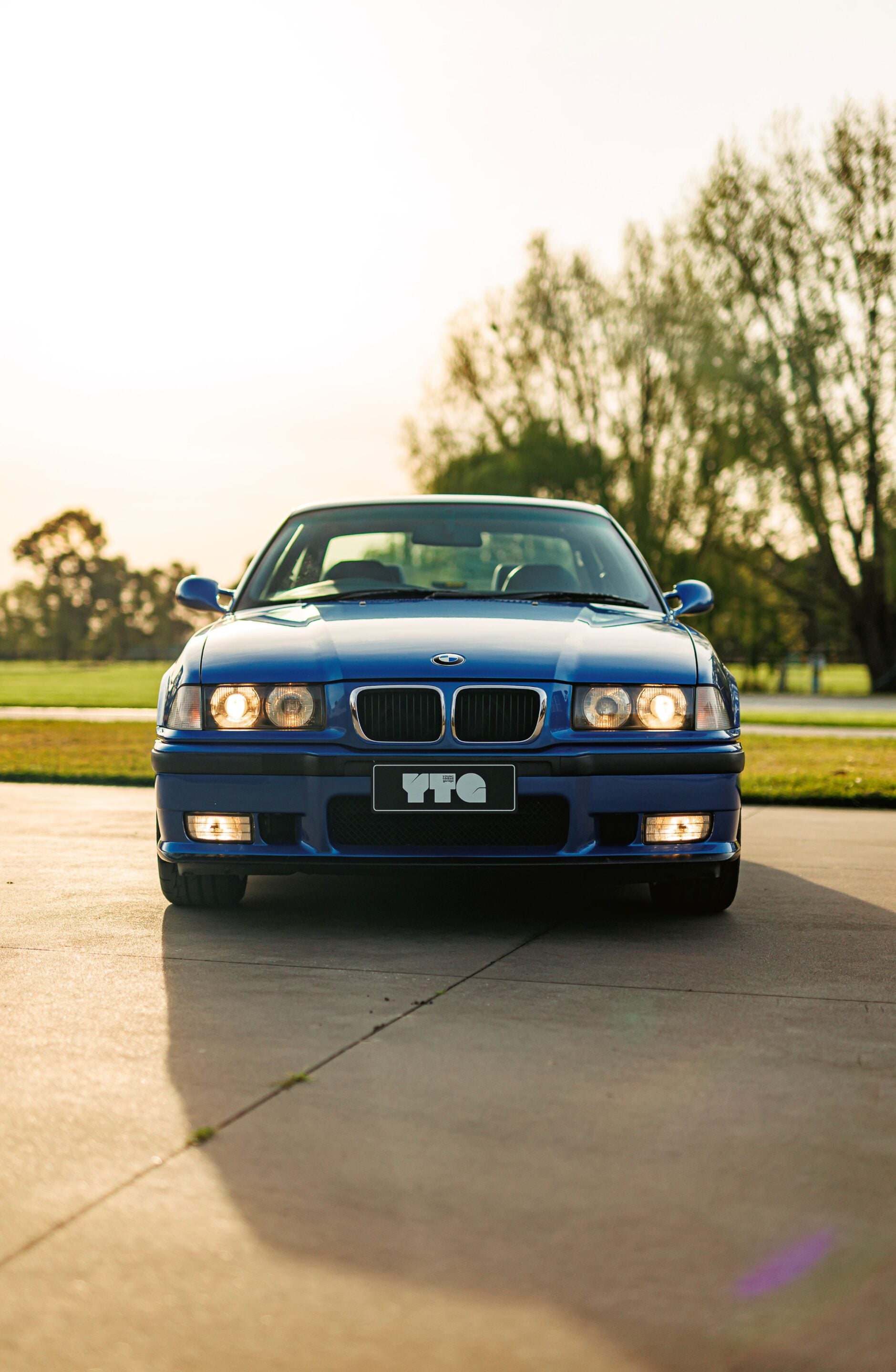 Blue BMW sedan facing the camera, headlights on, parked on a sunny driveway with trees in the background.