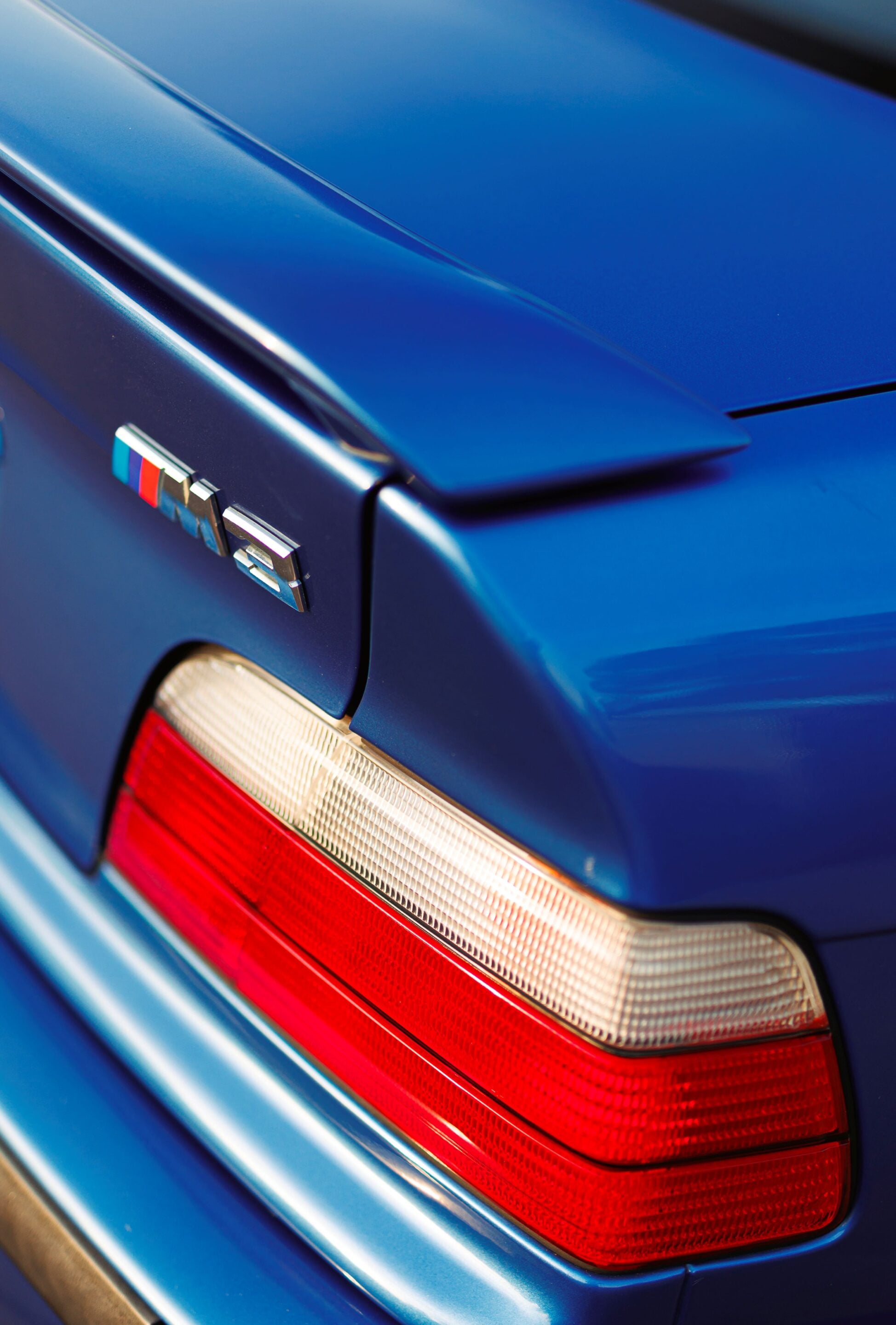 Close-up of a blue car's rear corner showing a red taillight and an M badge on the trunk edge.