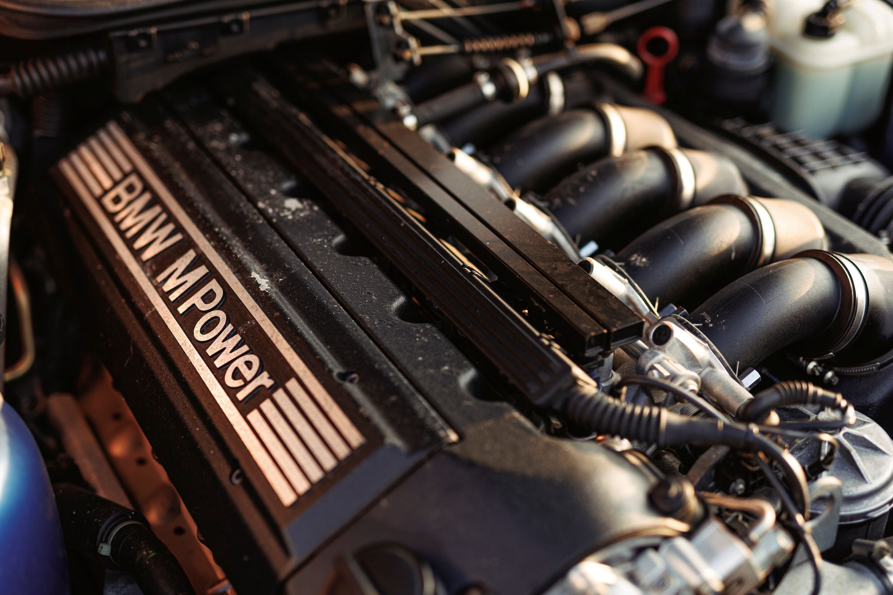 Close-up of a car engine bay featuring a BMW M Power valve cover with surrounding hoses and components.
