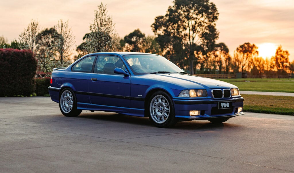 Blue BMW 3-series coupe parked on a concrete driveway at sunset with trees in the background.
