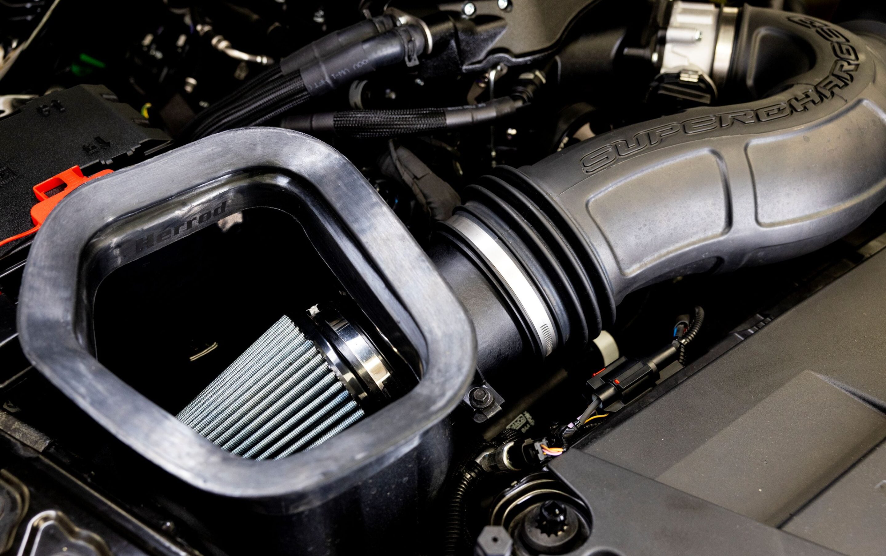 Close-up of a car engine bay showing a large air intake/air filter housing labeled 'Hartig' and a 'SUPERCHARGED' cover nearby (performance intake).