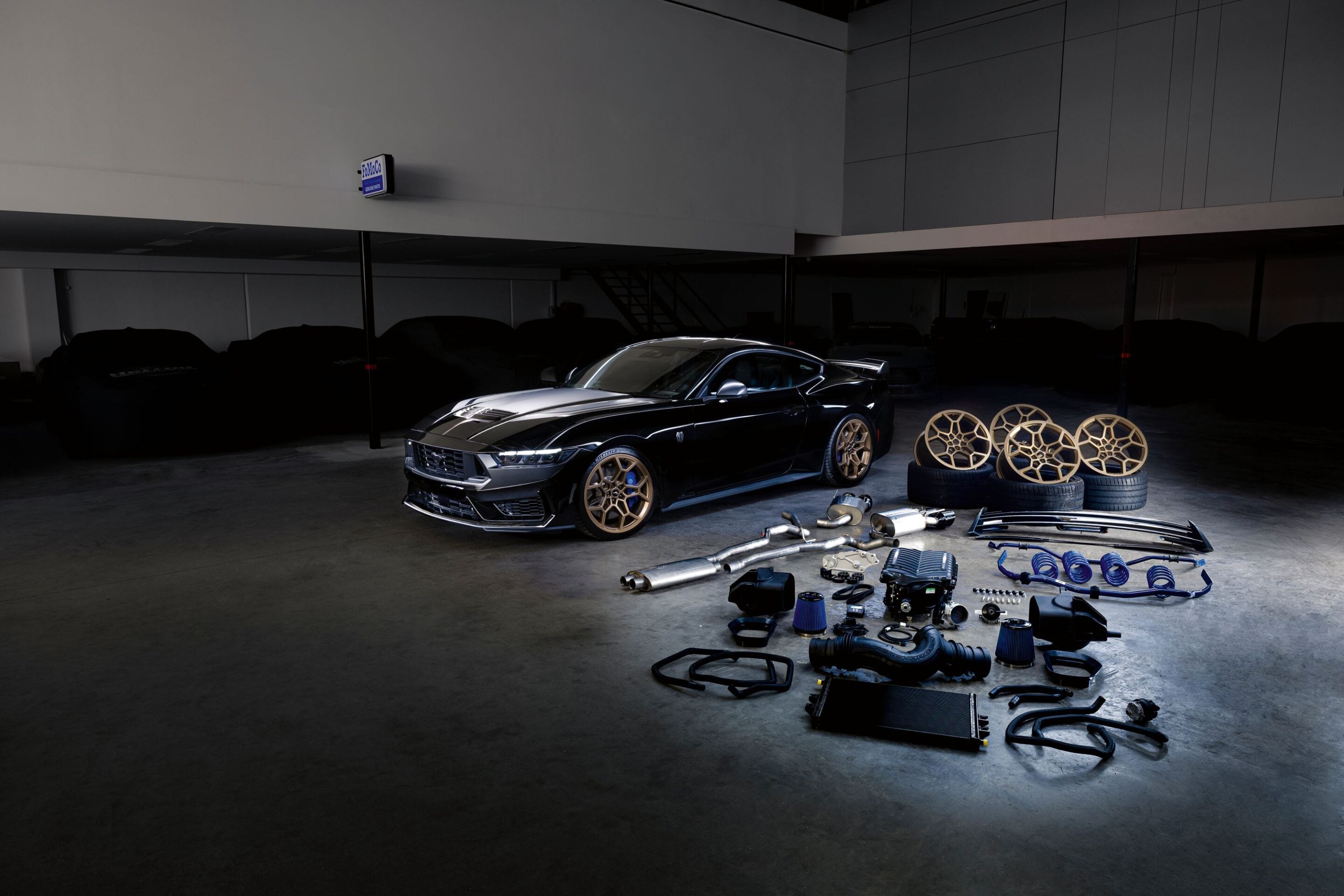 Black sports car in a dim garage surrounded by car parts and alloy wheels on the floor, foreground filled with loose components.