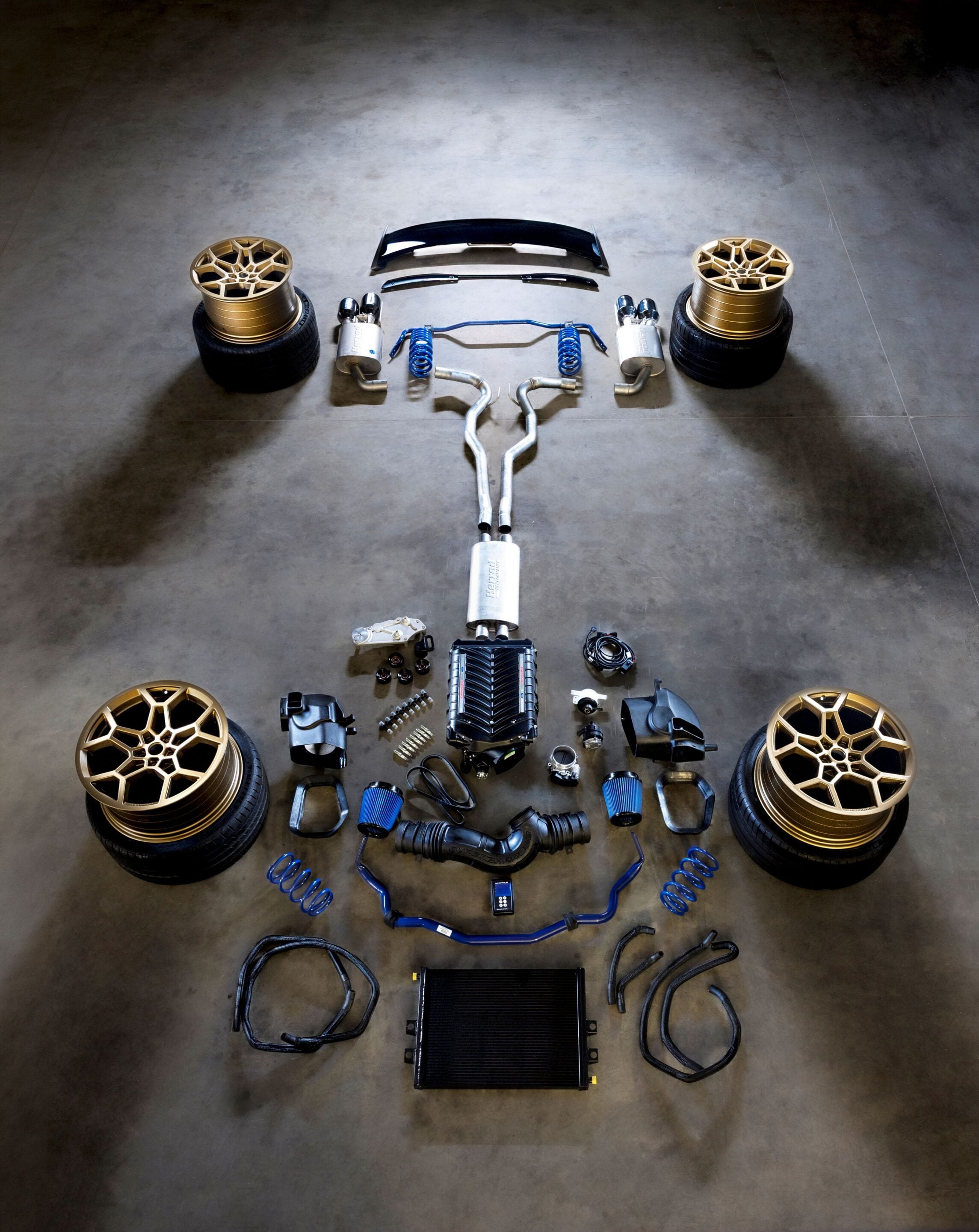 Disassembled car parts laid out in a symmetrical arrangement on a concrete floor: four gold alloy wheels with tires at corners, blue coilovers, exhausts, a radiator, hoses, and miscellaneous components laid in rows beneath a central assembly.