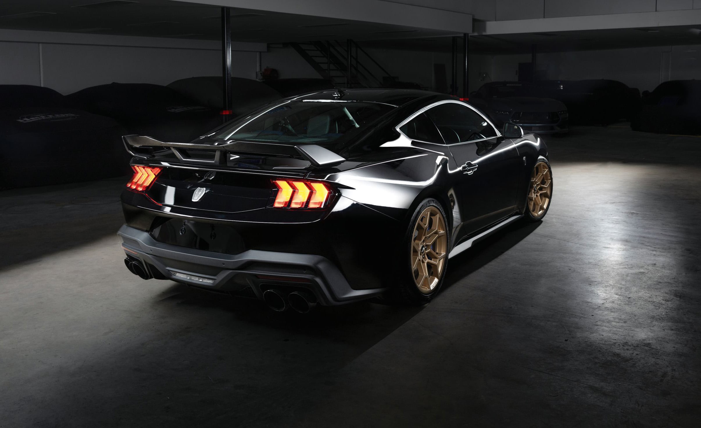 Black sports car with red taillights and gold wheels parked in a dim indoor garage, viewed from the rear side angle.