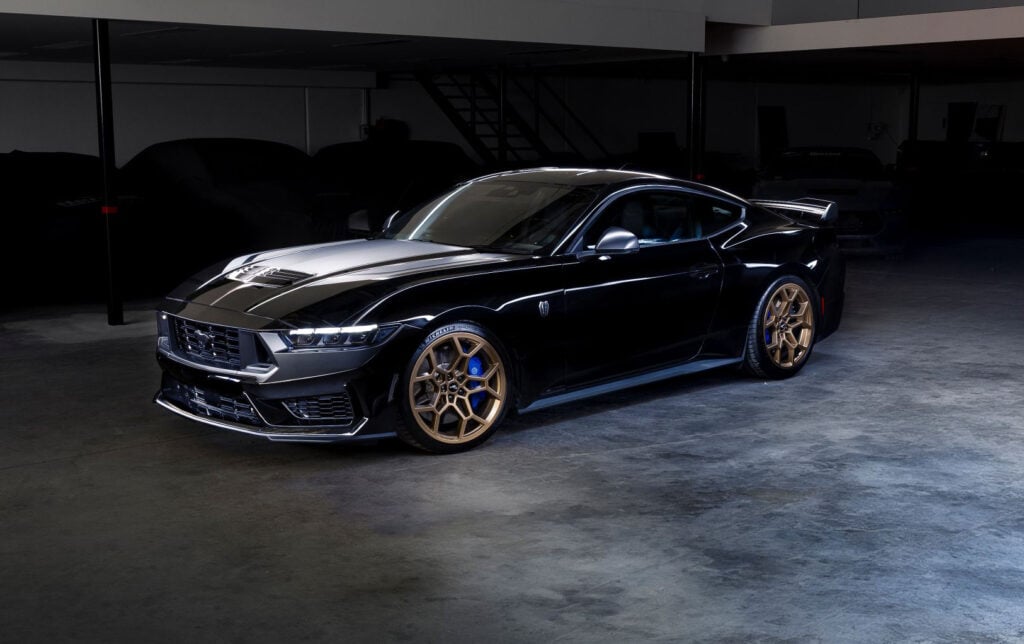 Front three-quarter view of a glossy black Ford Mustang with bronze/gold wheels in a dim showroom light.