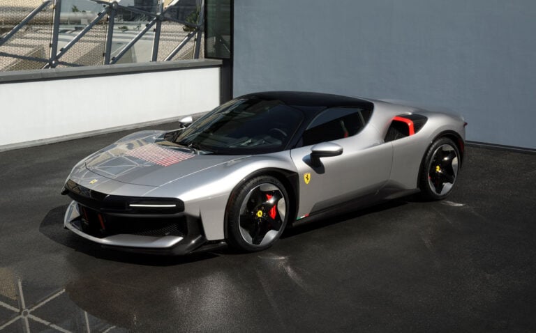 Silver Ferrari sports car in a three-quarter front view, showcasing red brake calipers, black roof, and sleek aerodynamic styling beside a blue-gray wall and glass railing.