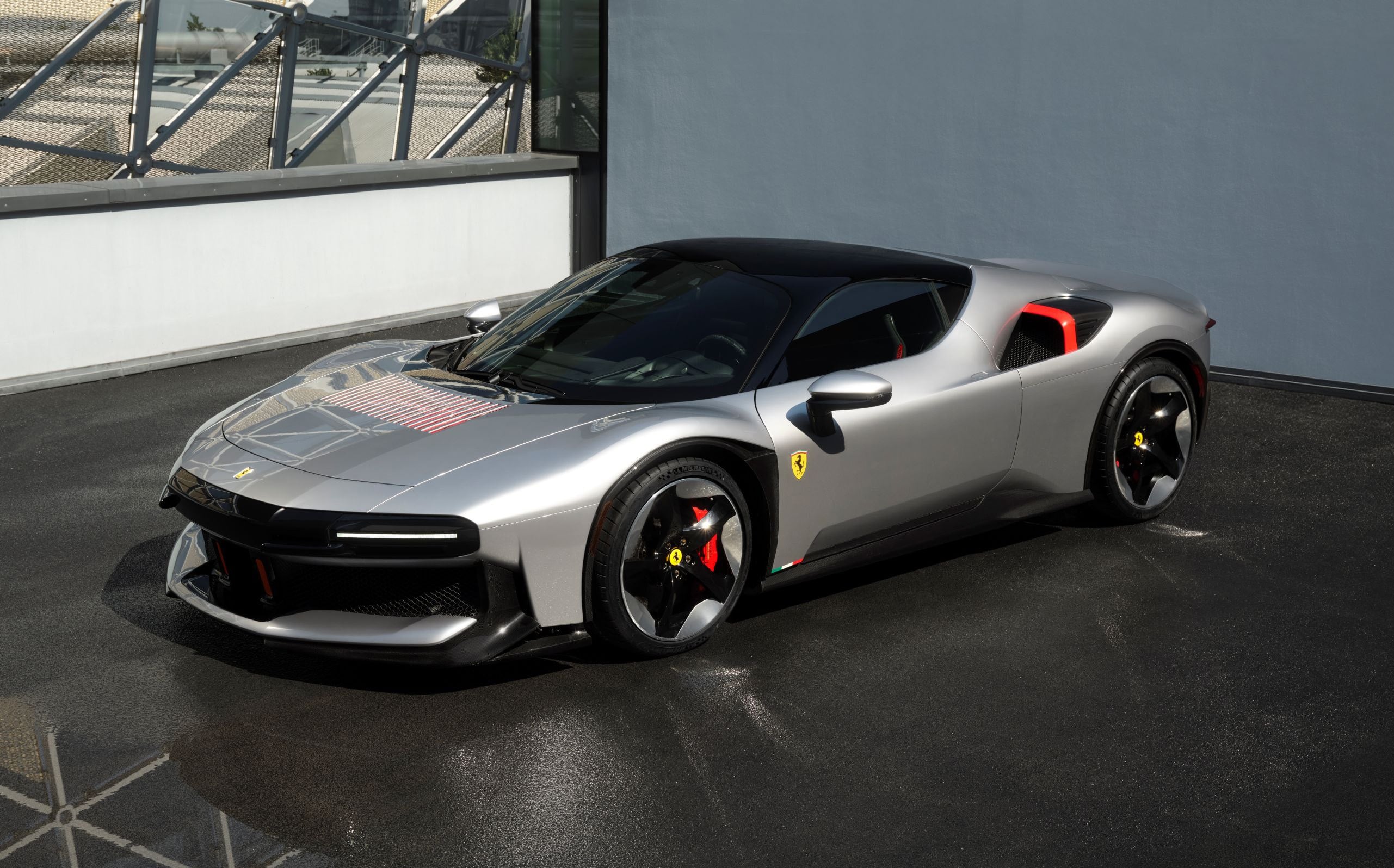 Silver Ferrari sports car in a three-quarter front view, showcasing red brake calipers, black roof, and sleek aerodynamic styling beside a blue-gray wall and glass railing.