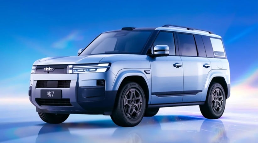 Three-quarter front view of a light blue SUV with roof rails, parked on a reflective surface under a blue sky.