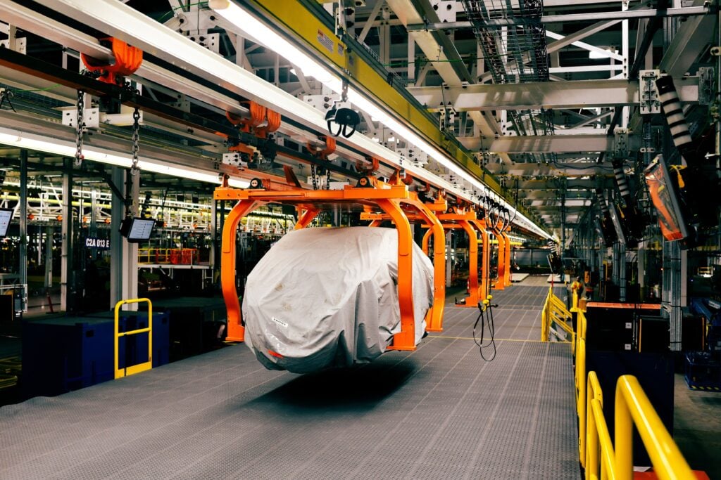 Industrial factory interior with an orange overhead hoist system carrying a large gray-wrapped load along a suspended track. Safety rails run along the walkway in the foreground.