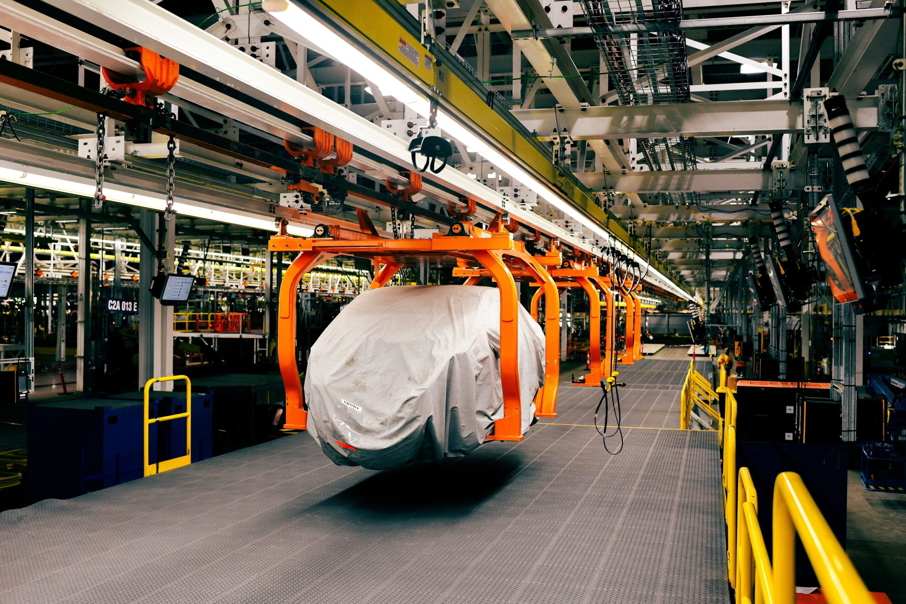 Industrial factory interior with an orange overhead hoist system carrying a large gray-wrapped load along a suspended track. Safety rails run along the walkway in the foreground.